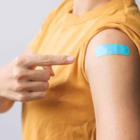 A woman points to her arm, where a blue bandage covers a vaccination injection site