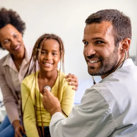 A pediatrician listens to a child's heartbeat with a parent in the background
