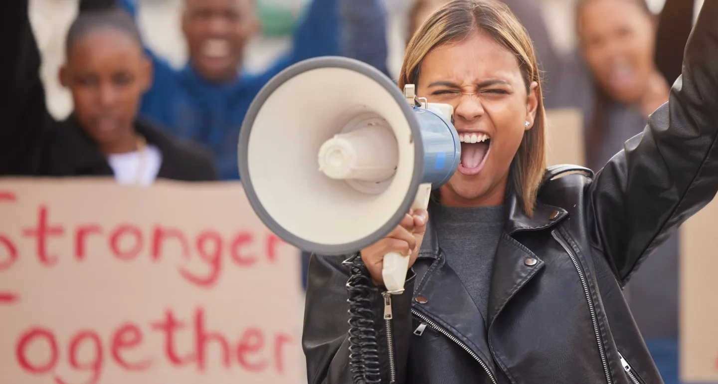 A woman with a raised fist cheers into a microphone; the crowd behind her holds a sign reading "Stronger Together"
