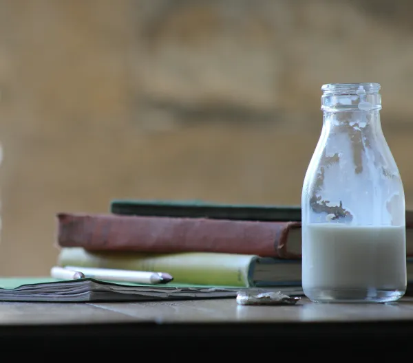 A small bottle of milk on a table beside textbooks and pencils