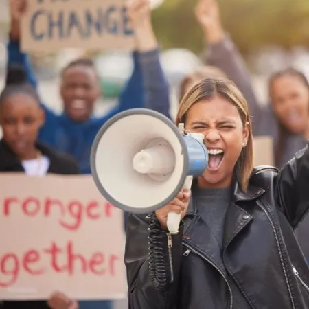A woman with a raised fist cheers into a microphone; the crowd behind her holds a sign reading "Stronger Together"